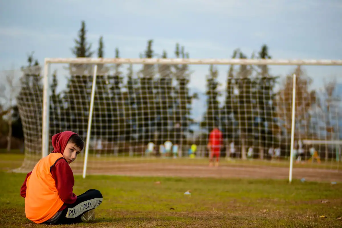 Băiat în echipament de fotbal stă jos lângă poarta unui teren de antrenament