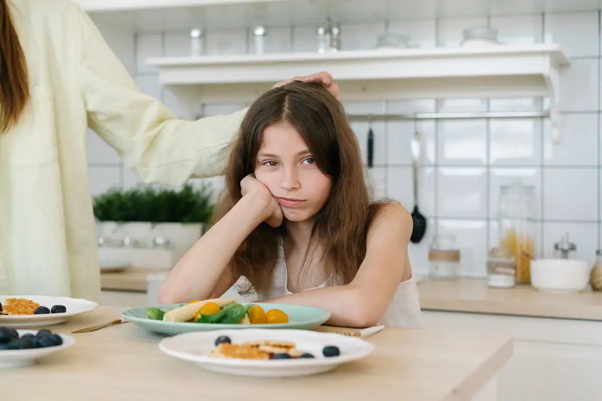 Fată cu mutră plictisită în fața unor farfurii cu fructe și legume