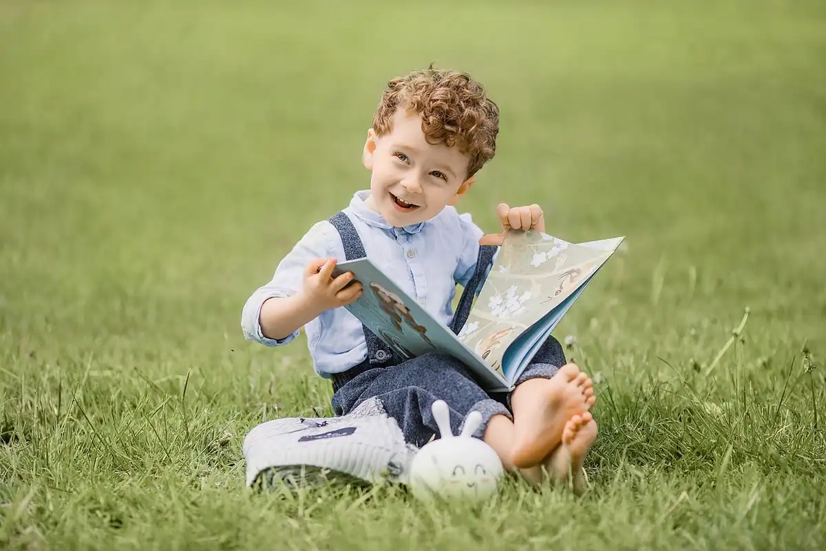 Băiețel așezat pe iarbă răsfoiește o carte ilustrată în parc
