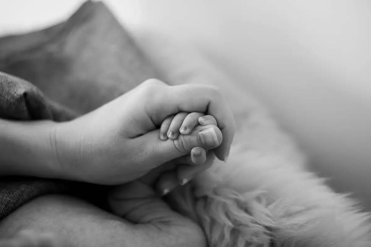 Mâna unui adult ține mânuța unui bebeluș, într-o fotografie alb-negru