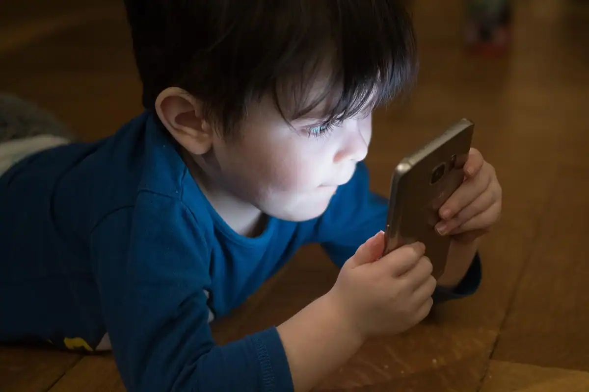 Băiețel întins pe podea se uită atent la un telefon mobil