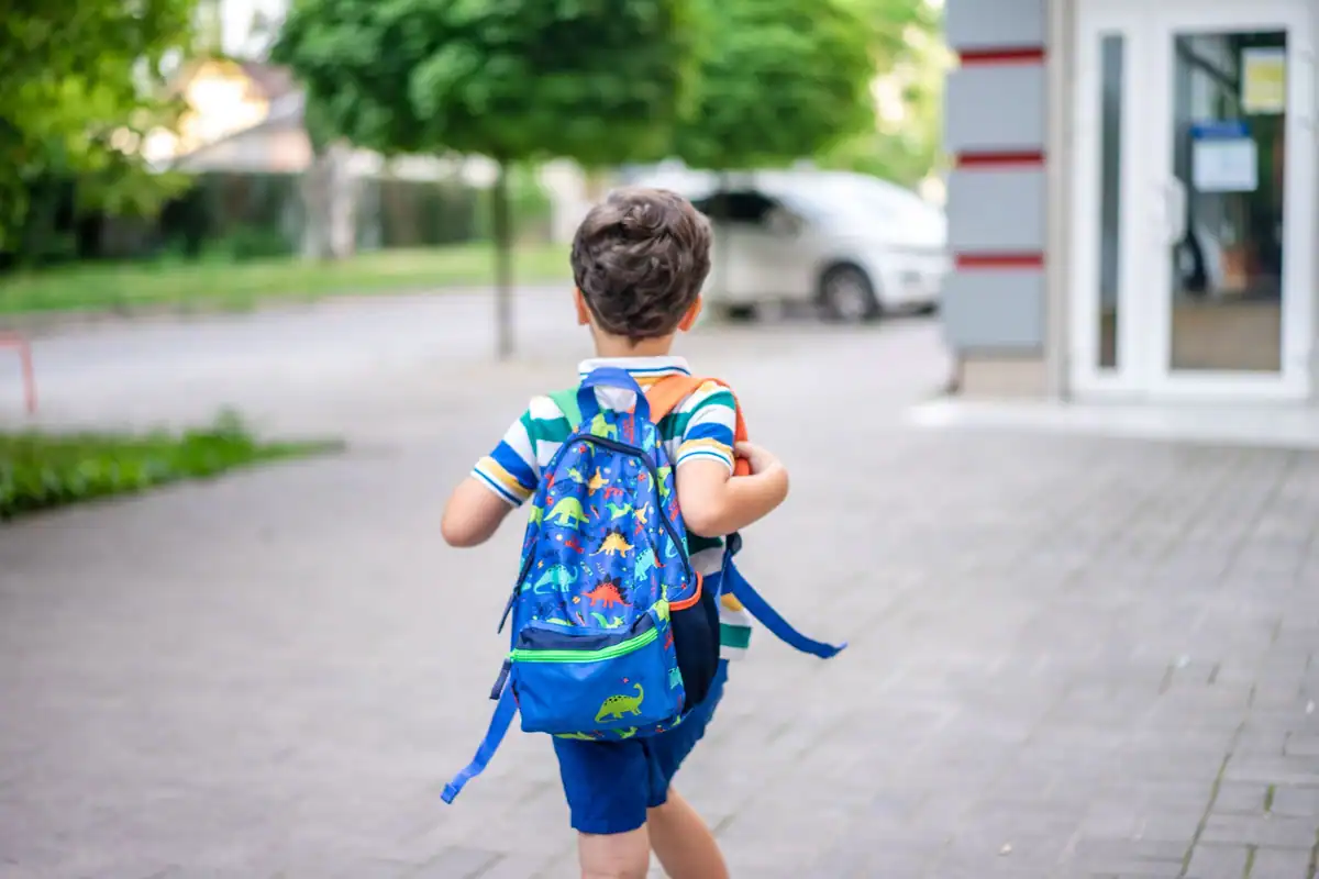 Copil cu un rucsac albastru merge pe trotuar spre școală, dimineața