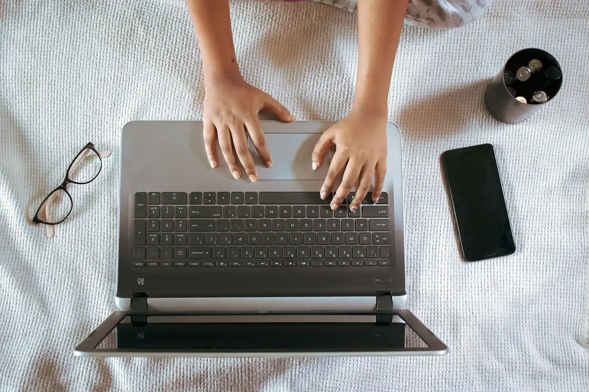 Mâini pe tastatura unui laptop așezat pe pat, lângă un telefon