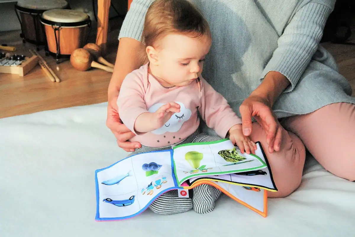 Bebeluș pe un cearșaf răsfoiește o carte cu poze, alături de mama